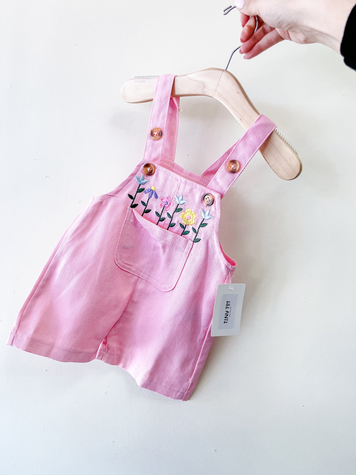 A hand holds the Tiny Tot Bundles Petal Patch Pink Overalls, featuring embroidered flowers on the front pocket, brown buttons, and a white tag attached to one strap.