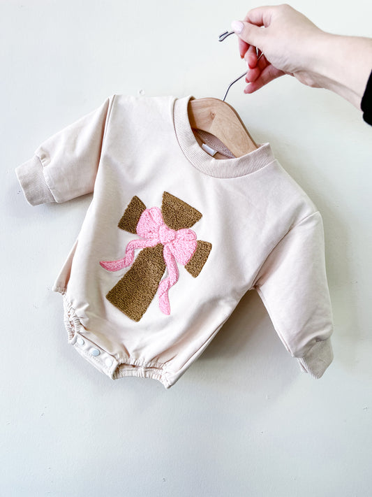 A hand holds the Tiny Tot Bundles Blush Blessings Cross Bubble, a light beige baby onesie with an embroidered brown cross and a large pink ribbon bow on the front, set against a plain, light-colored wall.