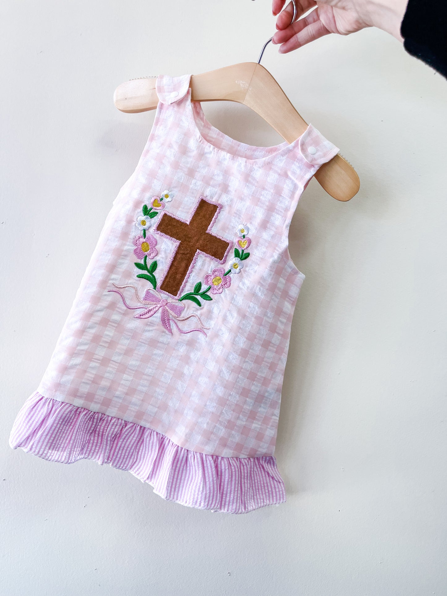 A hand holds a hanger with the Faith & Flowers Gingham Dress by Tiny Tot Bundles, featuring pink and white checks, a ruffled hem, and an embroidered brown cross surrounded by flowers on the front.