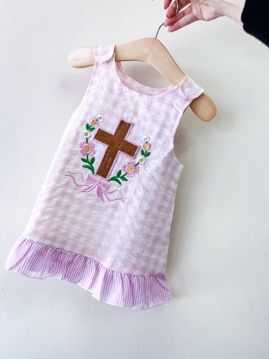 A hand holds a hanger with the Faith & Flowers Gingham Dress by Tiny Tot Bundles, featuring pink and white checks, a ruffled hem, and an embroidered brown cross surrounded by flowers on the front.