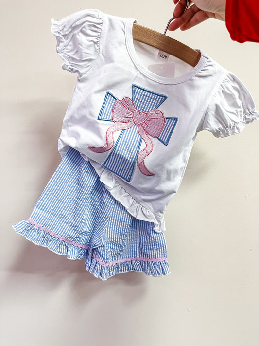 The Sunday Blessing Blue Set by Tiny Tot Bundles, featuring a white short-sleeve top with a blue cross and pink bow and blue-and-white striped ruffle shorts, is displayed on a hanger against a plain background.