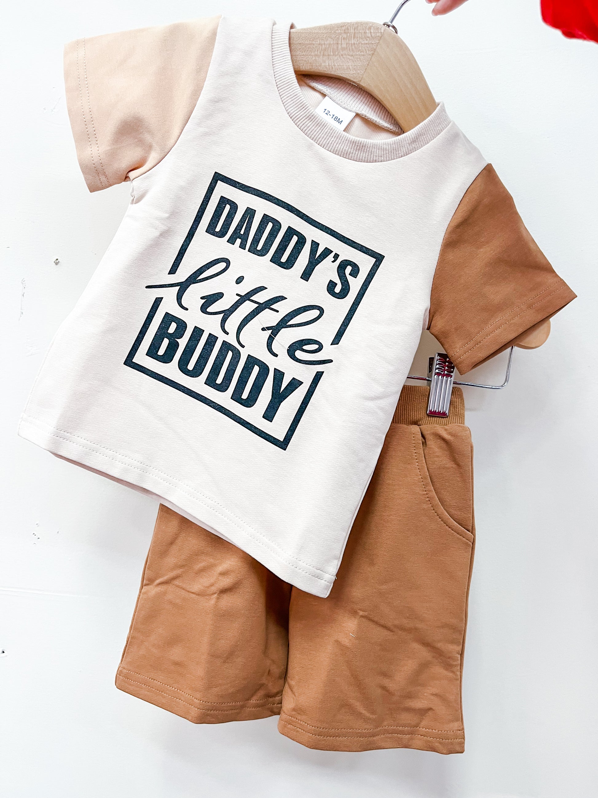 The Daddy's Little Buddy Short Set by Tiny Tot Bundles features a beige T-shirt with tan sleeves and "Daddy's little buddy" on the front, paired with matching tan shorts, displayed on a hanger.