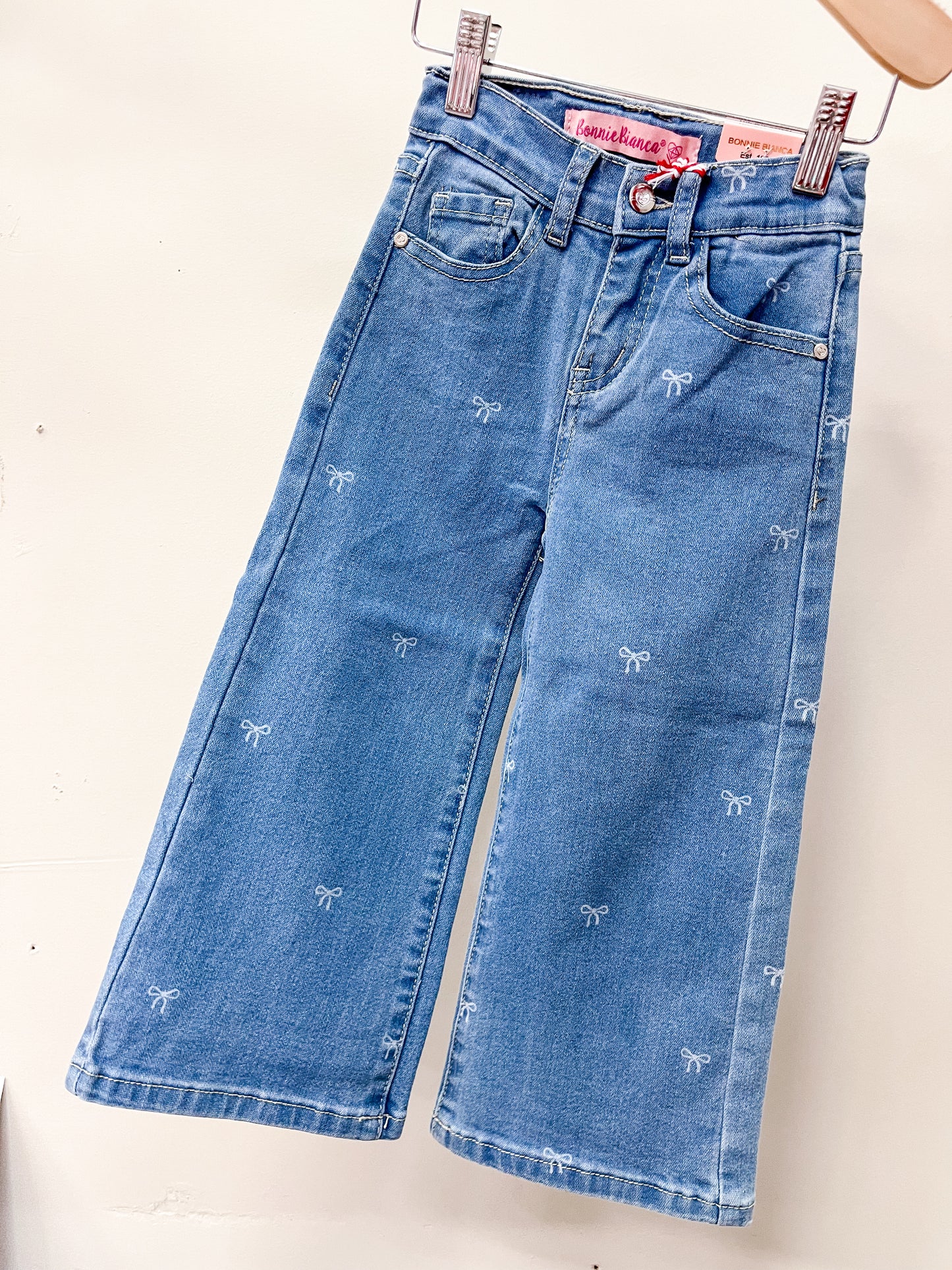 Cutie Patootie’s Bow Babe Denim Jeans feature a wide-leg cut, two front pockets, belt loops, and scattered white bow embroidery. Displayed on a hanger against a white background.