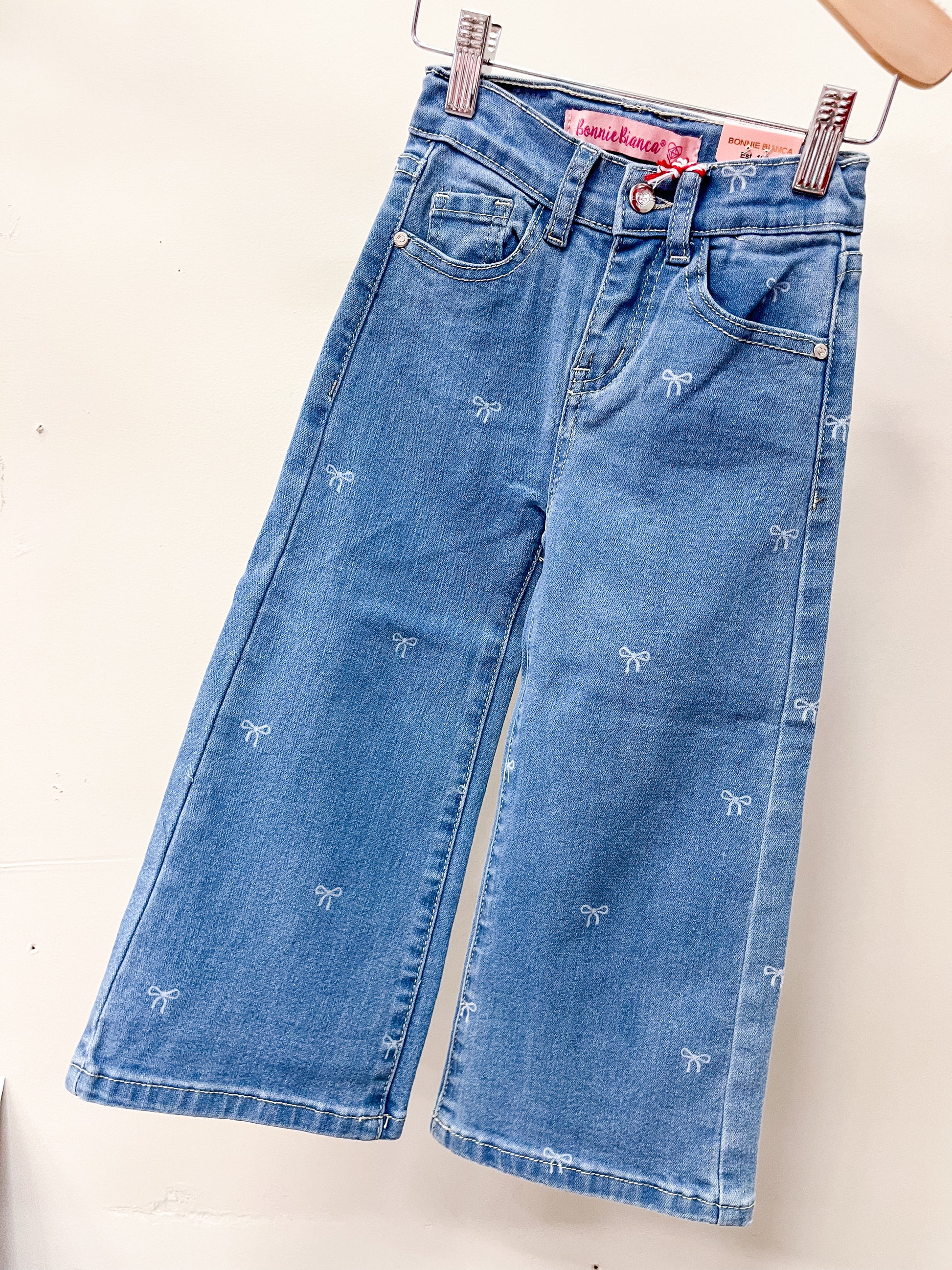 Cutie Patootie’s Bow Babe Denim Jeans feature a wide-leg cut, two front pockets, belt loops, and scattered white bow embroidery. Displayed on a hanger against a white background.