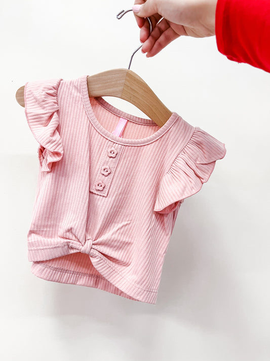 A hand holds a Cutie Patootie Rosewood Flutter Ribbed Top—a pink, short-sleeved baby shirt with ruffled sleeves, three front buttons, and a knotted hem—displayed on a wooden hanger.
