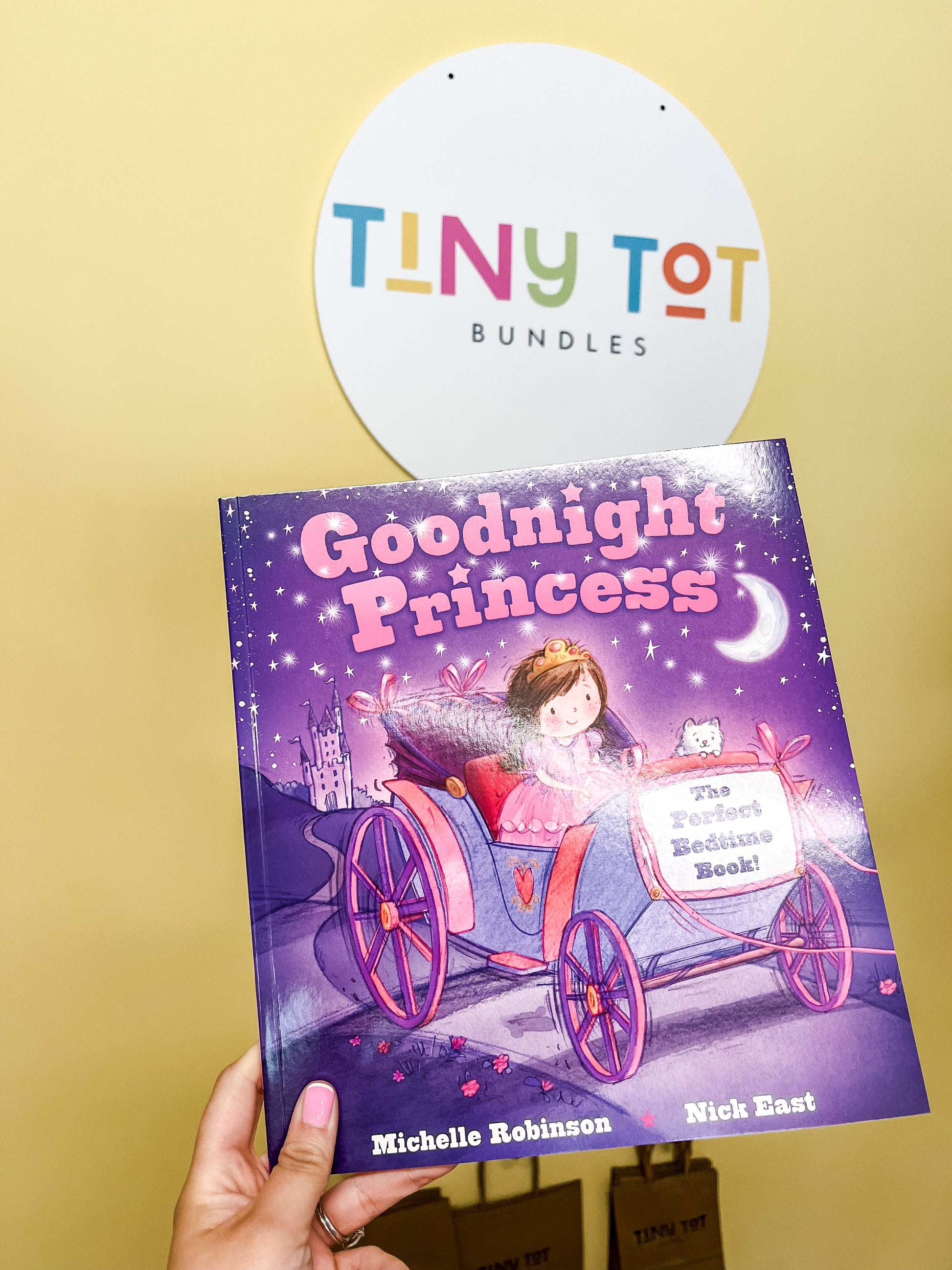 A hand holds the Goodnight Princess Book by Tiny Tot Bundles in front of a circular TINY TOT BUNDLES sign on a yellow wall. The cover features a princess in a pink carriage at night.