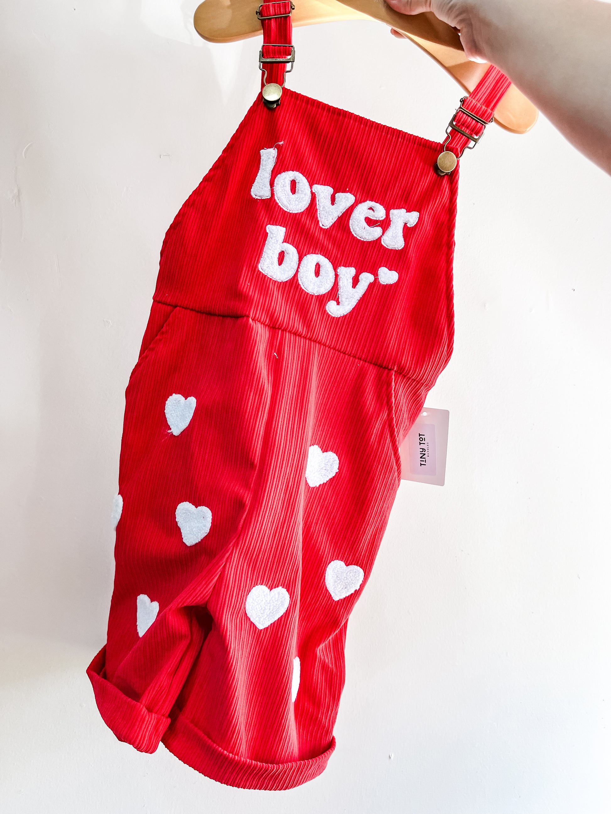 Tiny Tot Bundles' Lover Boy Red Overalls, featuring white hearts and "lover boy" on the front, are shown held up by hand against a plain white background.