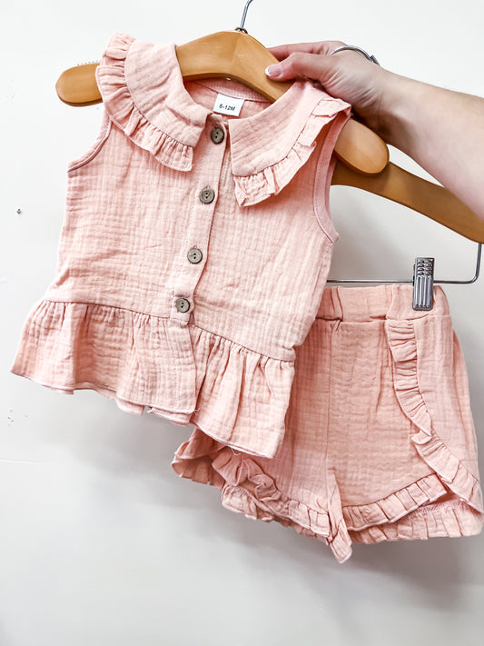 A hand holds the Tiny Tot Bundles Peachy Picnic Set: a matching pink toddler outfit with a sleeveless ruffled button-up top and elastic-waist ruffle shorts, both crafted from soft, textured fabric.