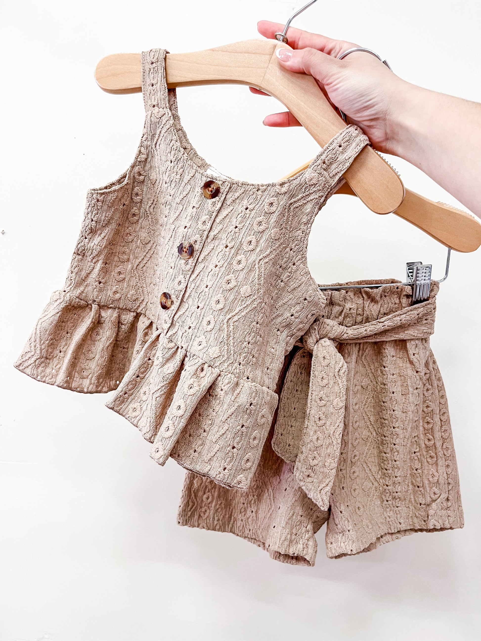 A hand holds the Cocoa Ruffle Set by Tiny Tot Bundles: a matching beige toddler outfit with a sleeveless ruffled top with buttons and textured design, plus high-waisted shorts with a fabric belt, both in lightweight material.