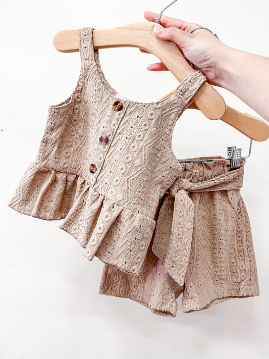 A hand holds the Cocoa Ruffle Set by Tiny Tot Bundles: a matching beige toddler outfit with a sleeveless ruffled top with buttons and textured design, plus high-waisted shorts with a fabric belt, both in lightweight material.