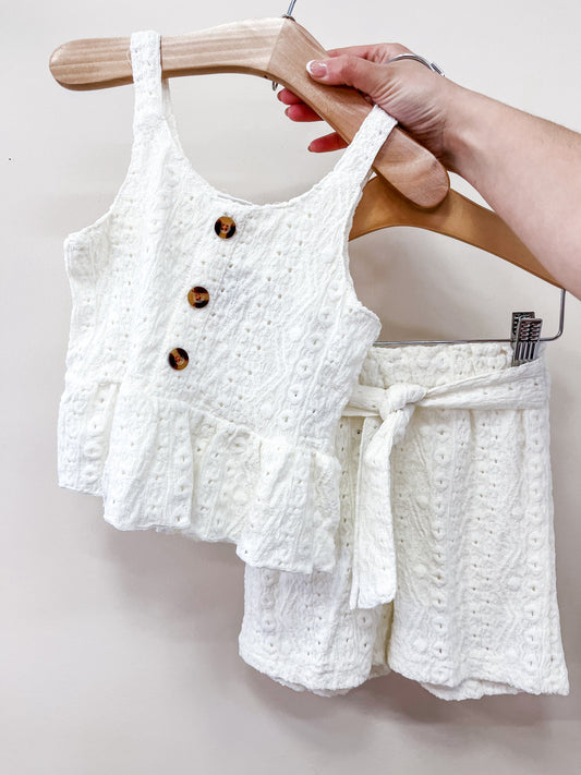 A hand holds a Tiny Tot Bundles Sweet Cream Tank Set: a sleeveless ruffled white top with three brown buttons and matching shorts with a tie belt, displayed on a wooden hanger against a light background.