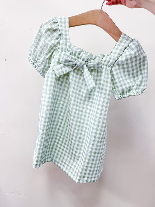 A hand holds a hanger displaying the Mint Picnic Dress by Tiny Tot Bundles, featuring green and white gingham checks, short puffed sleeves, and a front bow.