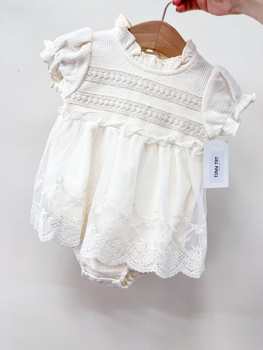 The Heirloom Lace Bubble by Tiny Tot Bundles is a cream baby romper with lace details, puffed short sleeves, and embroidered florals. Shown on a wooden hanger against a white background, held by hand, with a white tag attached.