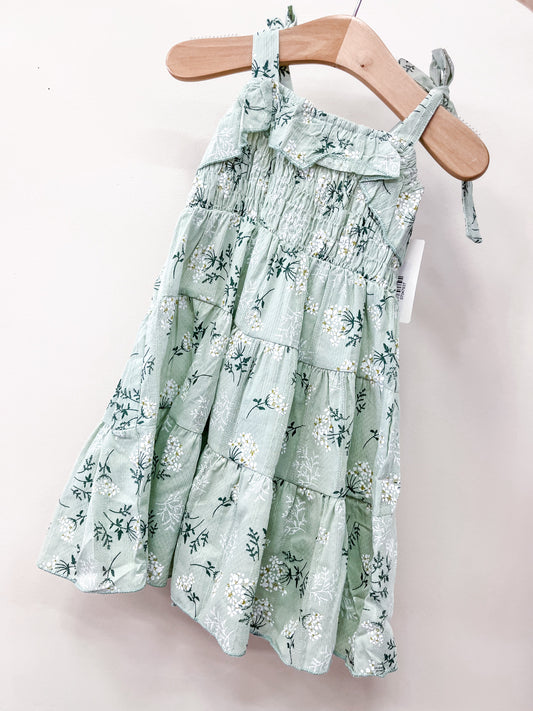 The Tiny Tot Bundles Sage Mellow Dress is a young girl's light green, sleeveless sundress with floral print, ruffled top edge, tiered design, and adjustable shoulder ties, shown hanging on a wooden hanger.