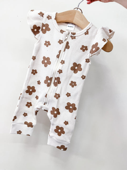 The Blush Daisy Zippy by Tiny Tot Bundles, a white baby romper with short ruffled sleeves, brown daisy prints, and a front zipper, is displayed on a wooden hanger against a plain white background.