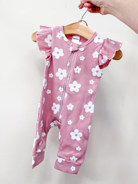 A hand holds the Tiny Tot Bundles Blush Daisy Zippy, a pink sleeveless baby romper with white daisy patterns and ruffled shoulders, displayed against a plain white background.