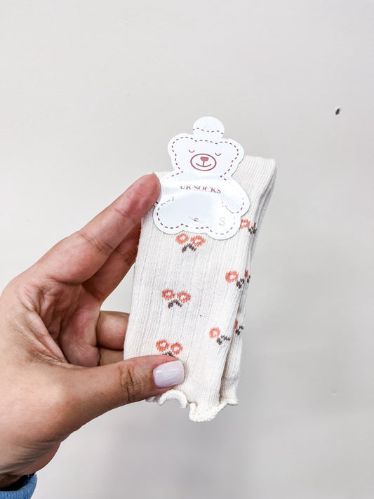 A hand holds Tiny Tot Bundles' Buttercream Daisy Socks—small, cream-colored socks with a floral pattern and scalloped edges on a bear-shaped label—against a white background.