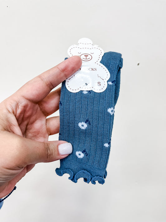 A hand with light-colored nail polish holds Tiny Tot Bundles' Dusty Blue Bloom Socks—small, blue ribbed socks with white polka dots, ruffled edges, and a bear-shaped tag attached at the top.