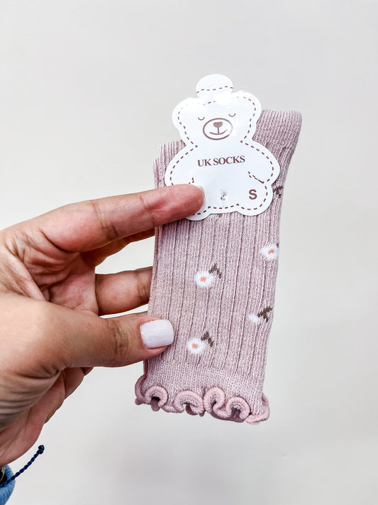 A hand with light pink nail polish holds a pair of Tiny Tot Bundles Blush Petal Socks, featuring a daisy pattern and ruffled edges. The pink baby socks are attached to a bear-shaped tag.