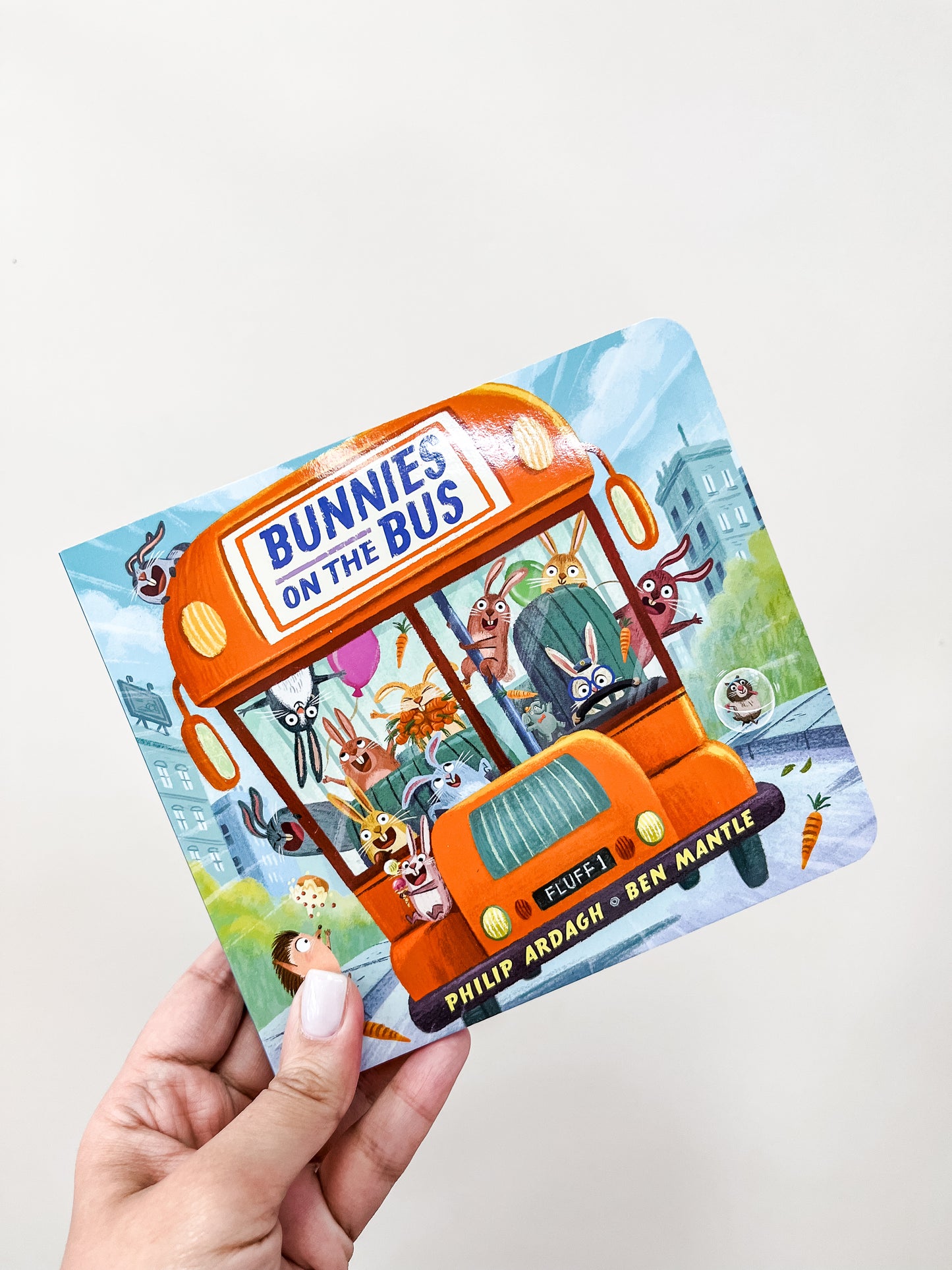 A hand holds the “Bunnies On the Bus Book” from Tiny Tot Bundles, showing illustrated bunnies riding a bright orange bus through a vibrant town.