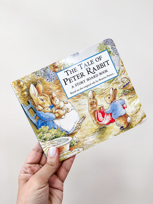 A hand holds the Tiny Tot Bundles Tale Peter Rabbit Story Book, showing an illustration of one adult and three young rabbits in clothes in a garden.