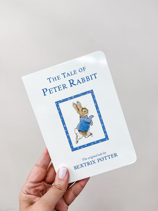 A hand holds the Tiny Tot Bundles "Tale of Peter The Rabbit Book Original," featuring Peter Rabbit in his blue coat on the white cover.
