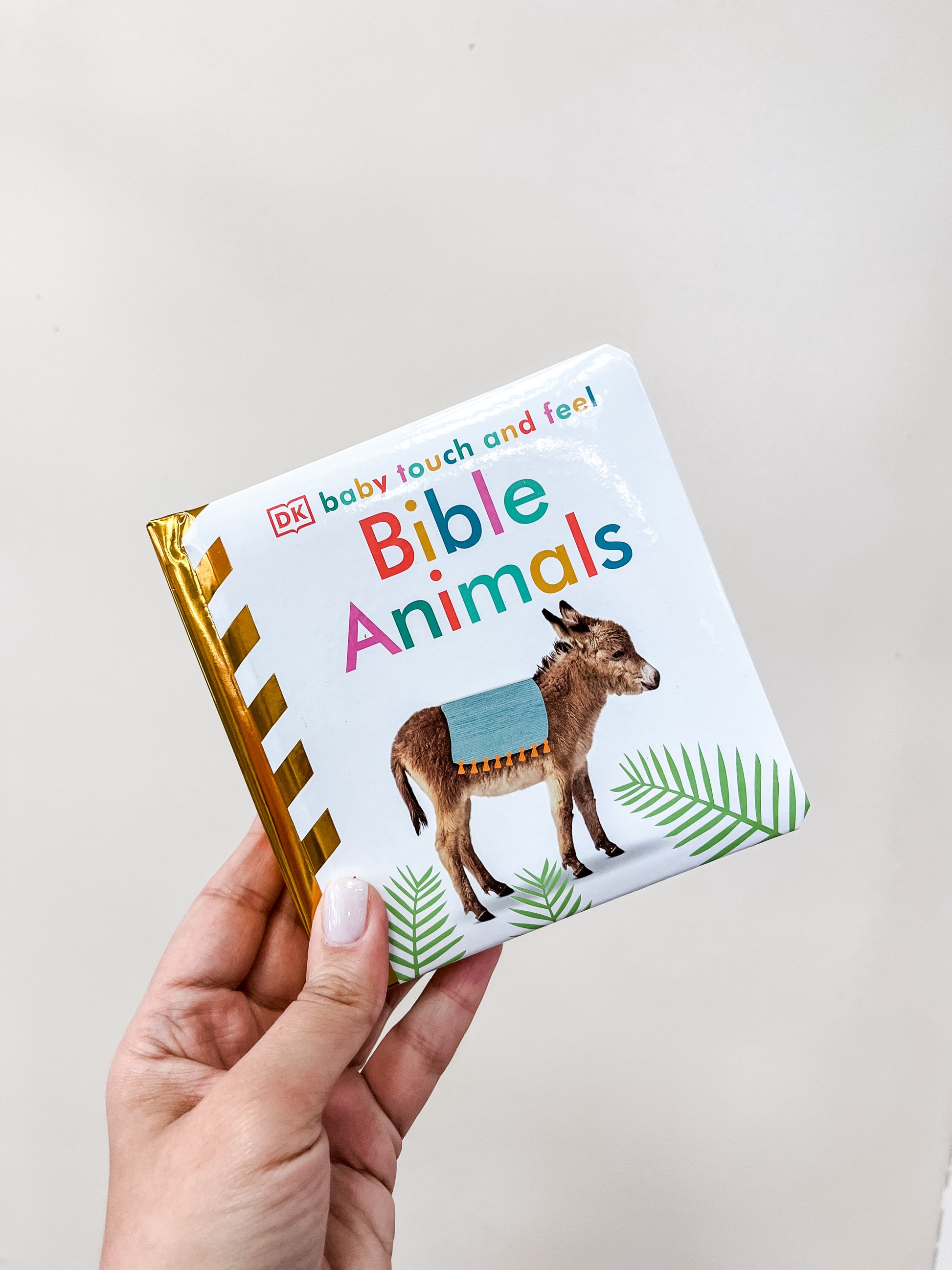 A hand holds the Bible Animals Baby Touch Book by Tiny Tot Bundles, featuring a donkey with a blue blanket on its back on the cover.