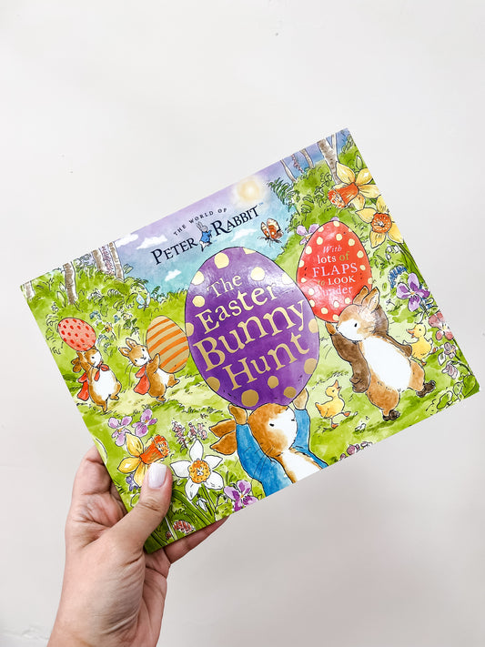 A hand holds the Peter The Rabbit Easter Bunny Hunt Book by Tiny Tot Bundles, featuring bunnies with decorated Easter eggs in a flower-filled garden on its colorful cover.