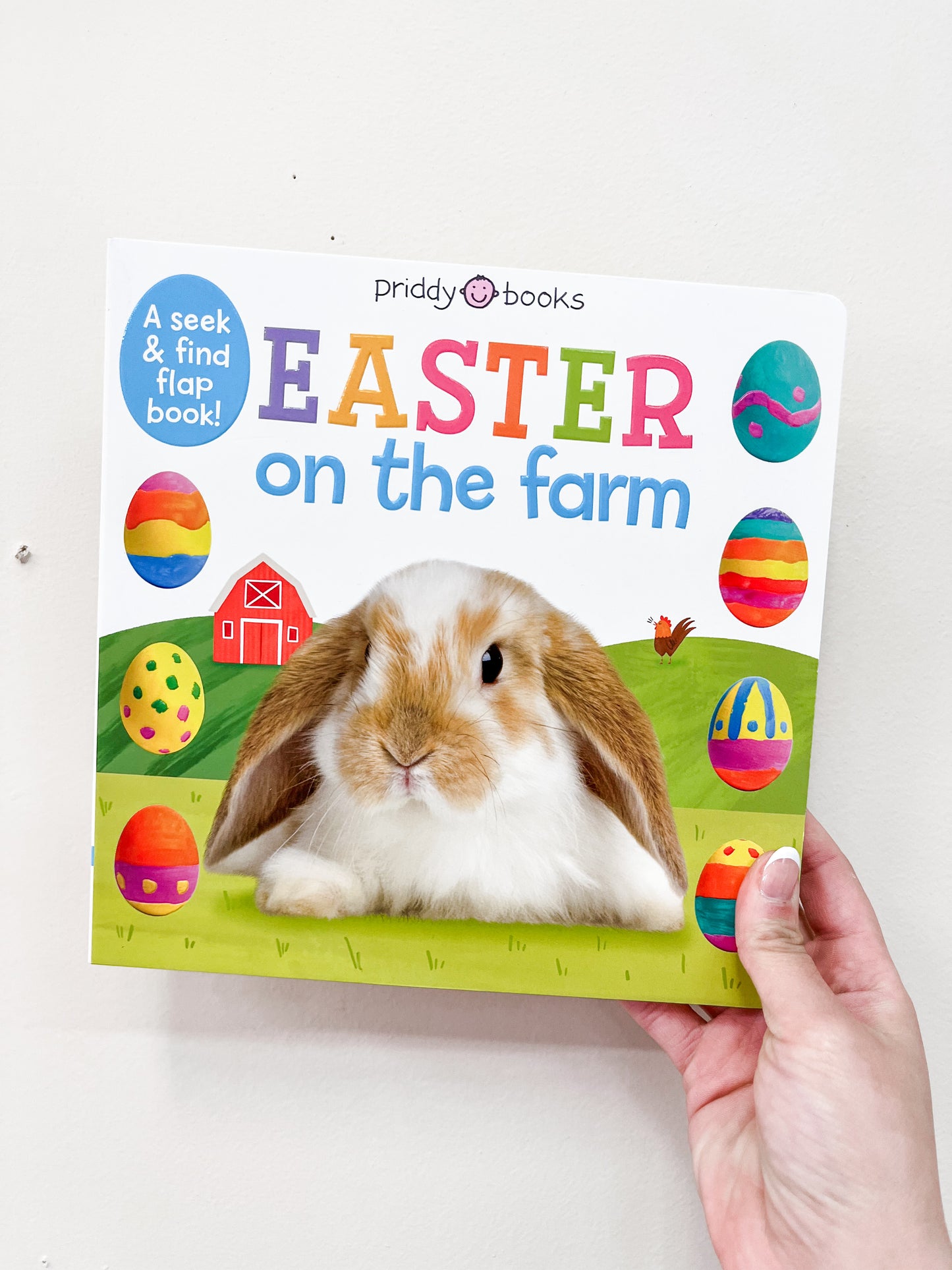 A hand holds the Easter On the Farm Flap Book by Macmillan Publishers, featuring a bunny in front of a barn with colorful Easter eggs on a white background.