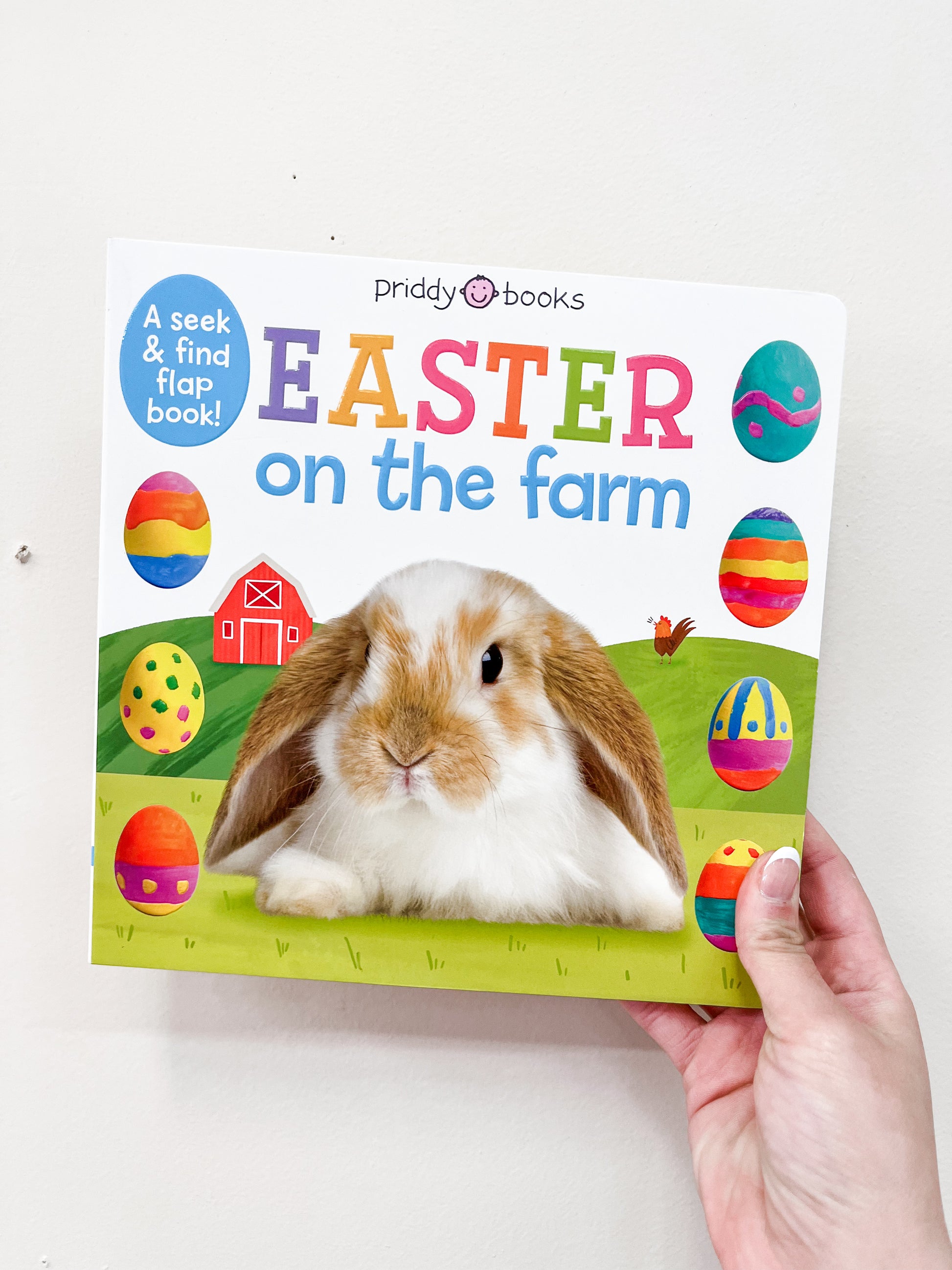 A hand holds the Easter On the Farm Flap Book by Macmillan Publishers, featuring a bunny in front of a barn with colorful Easter eggs on a white background.