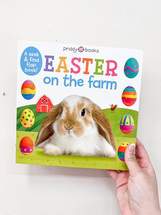 A hand holds the Easter On the Farm Flap Book by Macmillan Publishers, featuring a bunny in front of a barn with colorful Easter eggs on a white background.