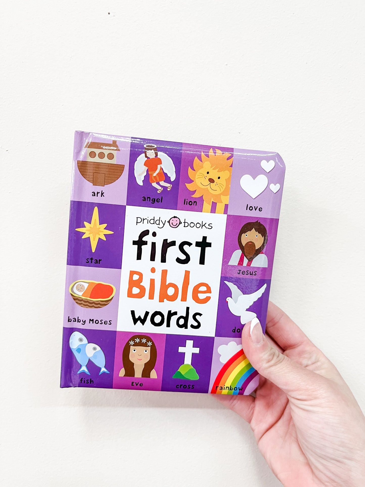 A hand holds the First Bible Words Book by Macmillan Publishers, featuring a colorful cover with biblical symbols and characters in purple, white, and orange.