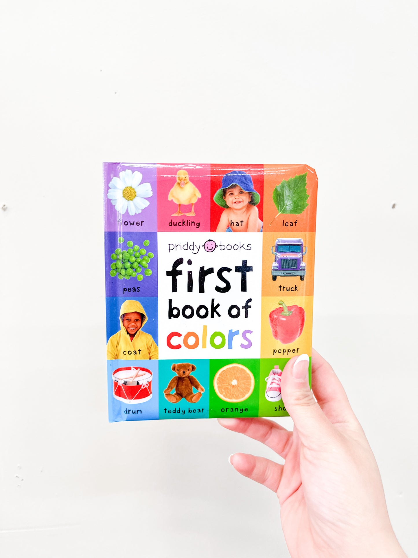 A hand holds the First Book of Colors Padded by Macmillan Publishers, a colorful children's book with cover images of a flower, duckling, hat, leaf, peas, coat, truck, pepper, drum, teddy bear, orange, and shoe.