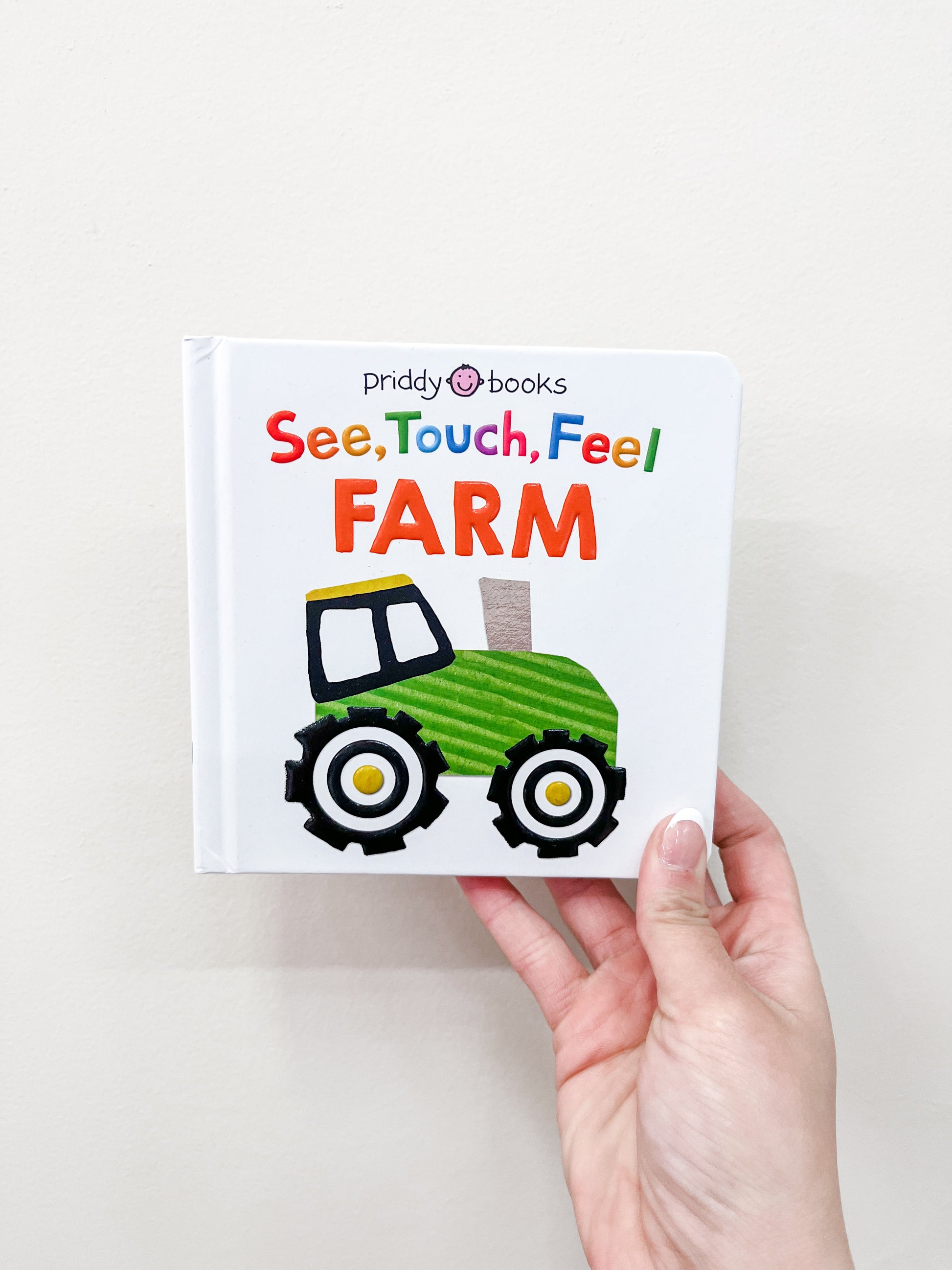 A hand holds the See Touch Feel - Farm Book by Tiny Tot Bundles, featuring a green tractor on the white cover.