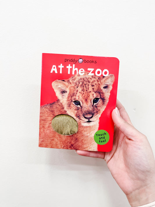 A hand holds the Macmillan Publishers "T & F - At the Zoo Book," a red children's book with a baby lion and furry touch-and-feel patch on the cover.