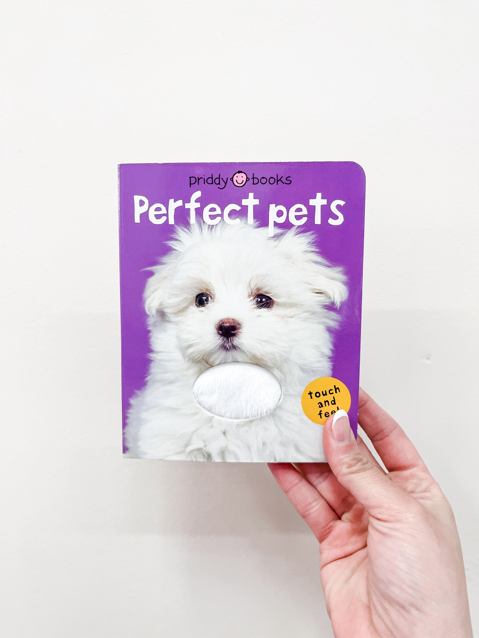 A hand holds the Macmillan Publishers T & F - Perfect Pets Book, featuring a fluffy white puppy on its purple cover and a textured patch for touching.