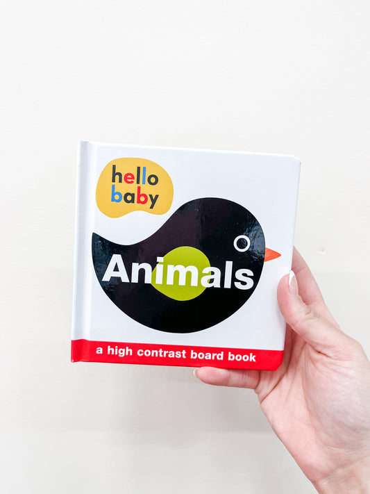 A hand holds the "Hello Baby Animals - Board Book" by Macmillan Publishers, featuring a cover with a black bird with a green wing and white eye on a white background.