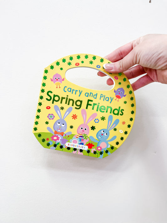 A hand holds a bunny-shaped children's book, Carry and Play: Spring Friends by Macmillan Publishers, featuring illustrated bunnies and birds on a yellow flower-decorated cover with a built-in handle.