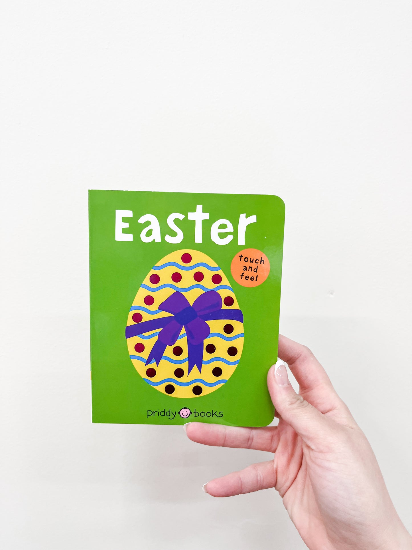 A hand holds the Touch and Feel Easter Book by Macmillan Publishers, a small green children's book with a colorful egg and purple bow on the cover. A round orange sticker reads "touch and feel.