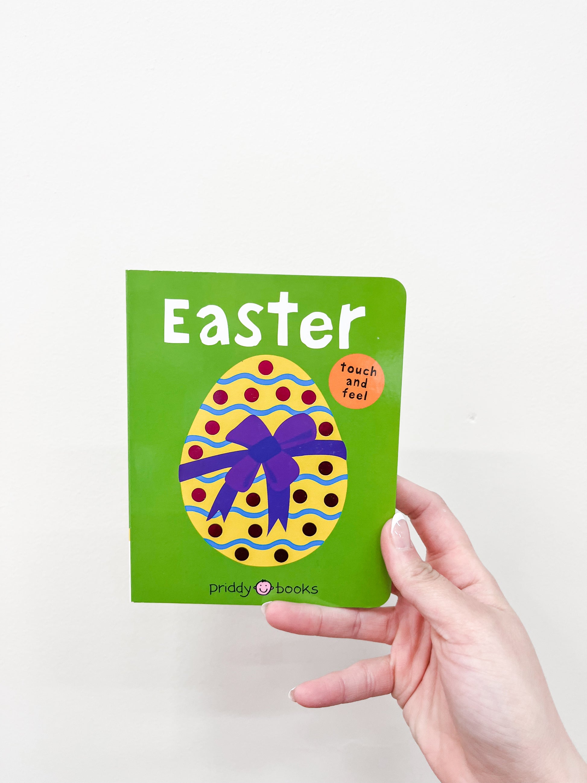 A hand holds the Touch and Feel Easter Book by Macmillan Publishers, a small green children's book with a colorful egg and purple bow on the cover. A round orange sticker reads "touch and feel.