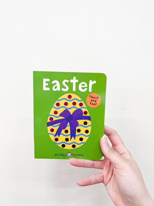A hand holds the Touch and Feel Easter Book by Macmillan Publishers, a small green children's book with a colorful egg and purple bow on the cover. A round orange sticker reads "touch and feel.