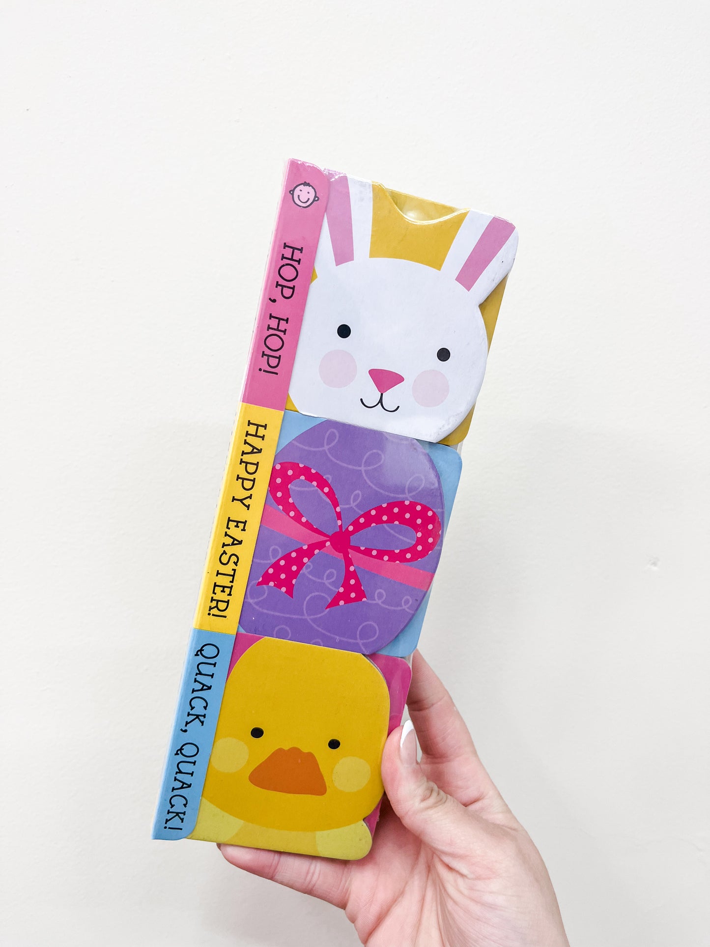 A hand holds Macmillan Publishers' "Chunky Pack: Easter: Hop-Hop!, Happy Easter!, Quack-Quack!" children's book, featuring illustrated bunny and chick faces on the cover and a pastel-colored spine with the titles.