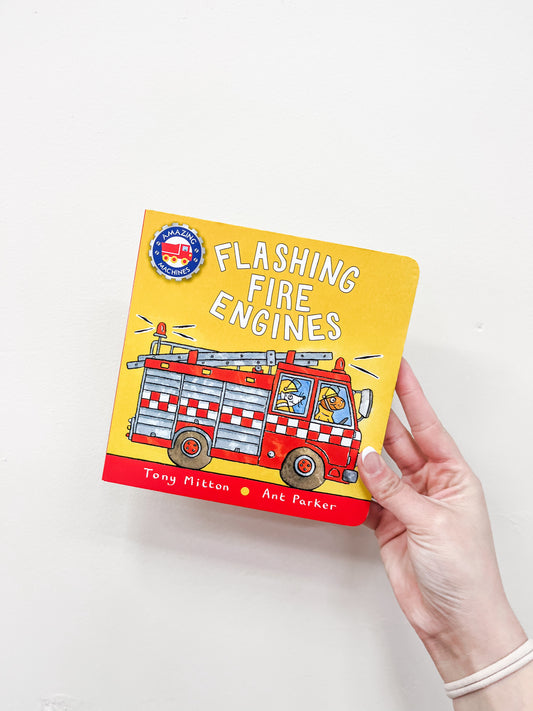 A hand holds the Macmillan Publishers' Flashing Fire Engines Book, featuring a yellow cover with a cartoon fire engine and two animal characters inside.