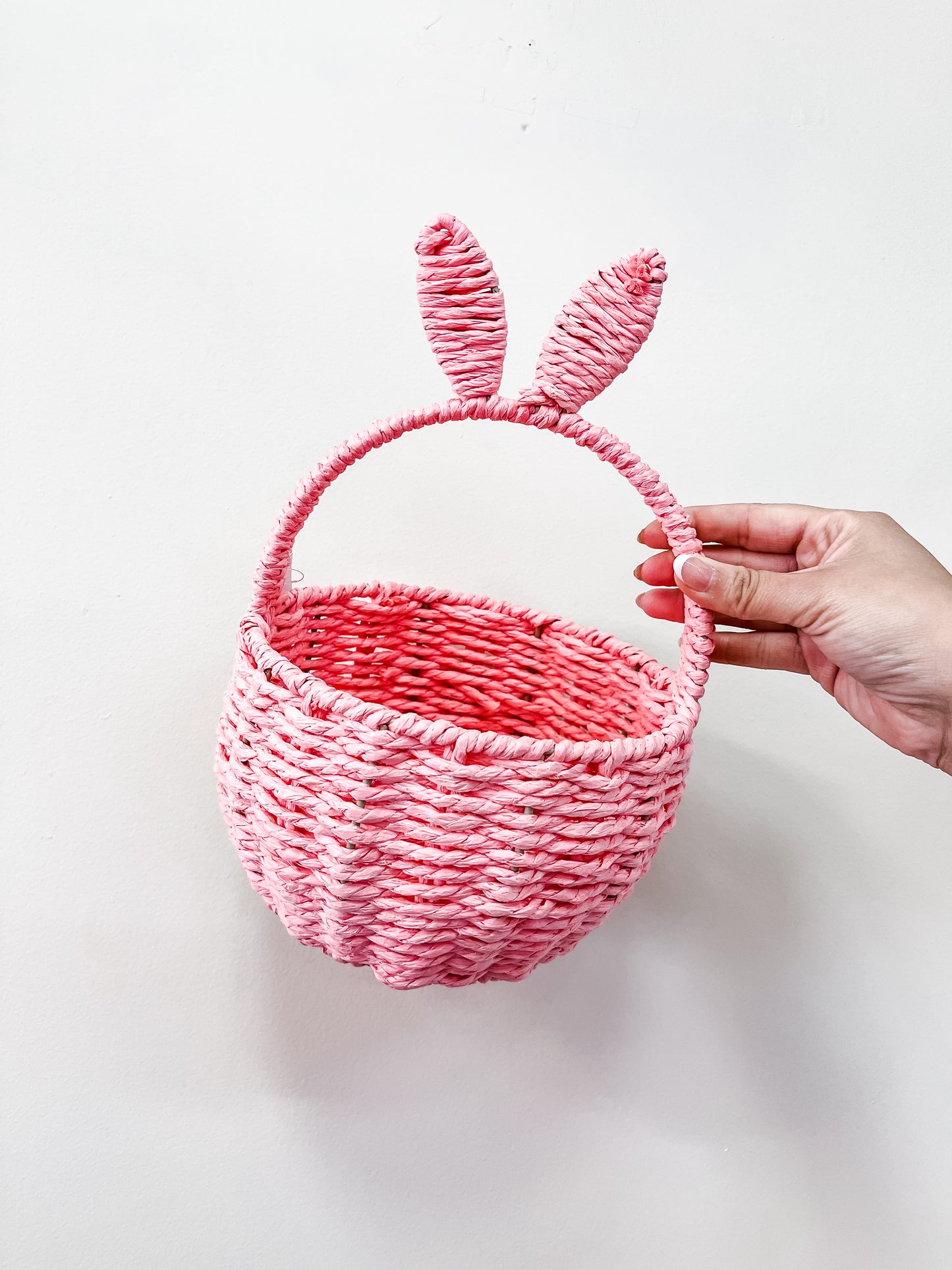 A hand holds the Capzone Pastel Bunny Ear Basket, a pink woven basket with two bunny ears on the handle, set against a plain white background.