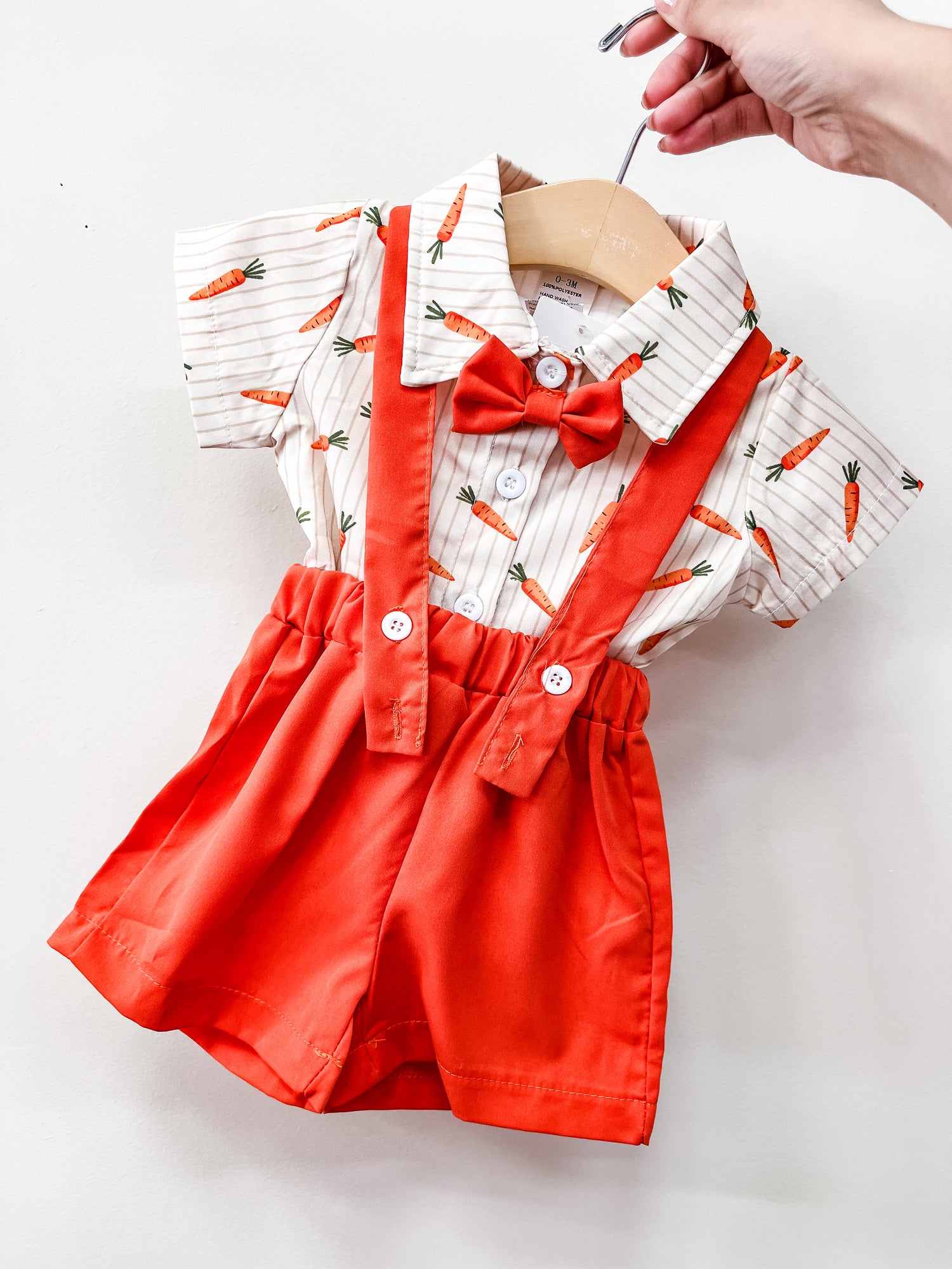 A hand holds the Tiny Tot Bundles Carrot Patch Suspender Set, featuring red suspenders, shorts, a matching bow tie, and a white shirt with orange carrots and thin stripes, displayed against a plain white background.