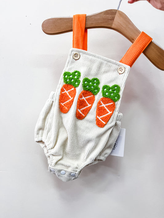 The Bunny Cord Overalls by Tiny Tot Bundles are cream-colored baby rompers with orange suspenders and three embroidered orange and green carrots on the front, shown hanging on a wooden hanger against a white background.