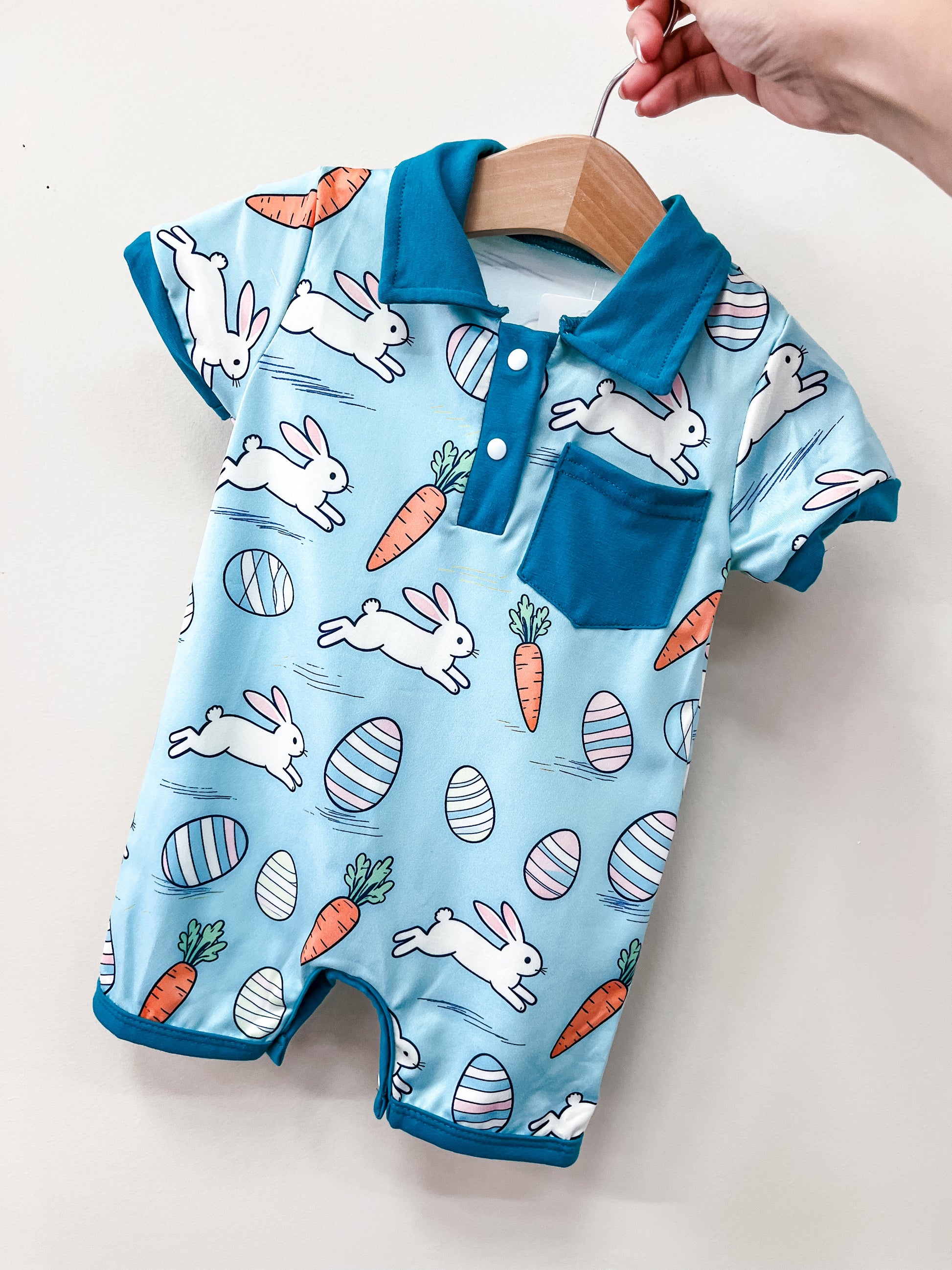 The Hoppy Day Polo Romper by Tiny Tot Bundles features a blue collar and pocket with playful white bunnies, carrots, and pastel Easter eggs. Displayed on a wooden hanger against a light background.