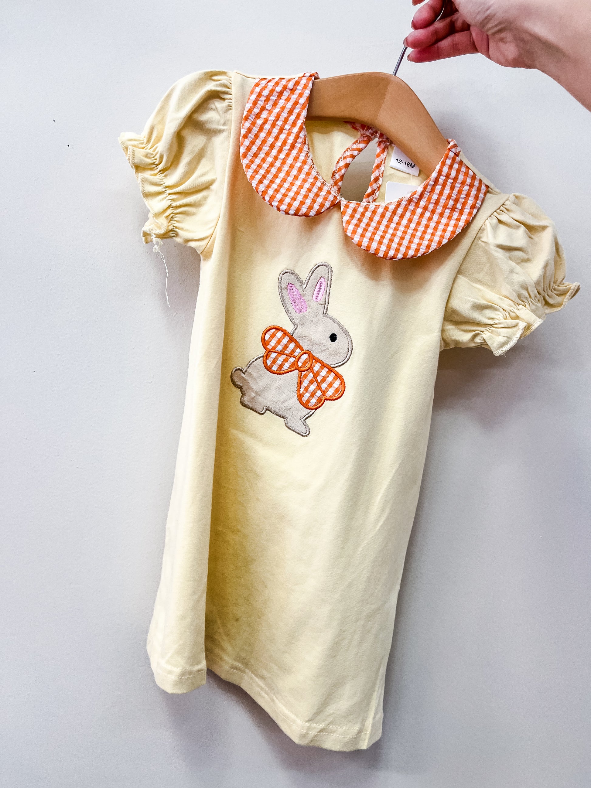 The Buttercup Bunny Dress by Tiny Tot Bundles is a yellow children’s dress with puffed sleeves, an orange and white gingham collar, and a bunny graphic with a matching bow tie, displayed on a hanger.
