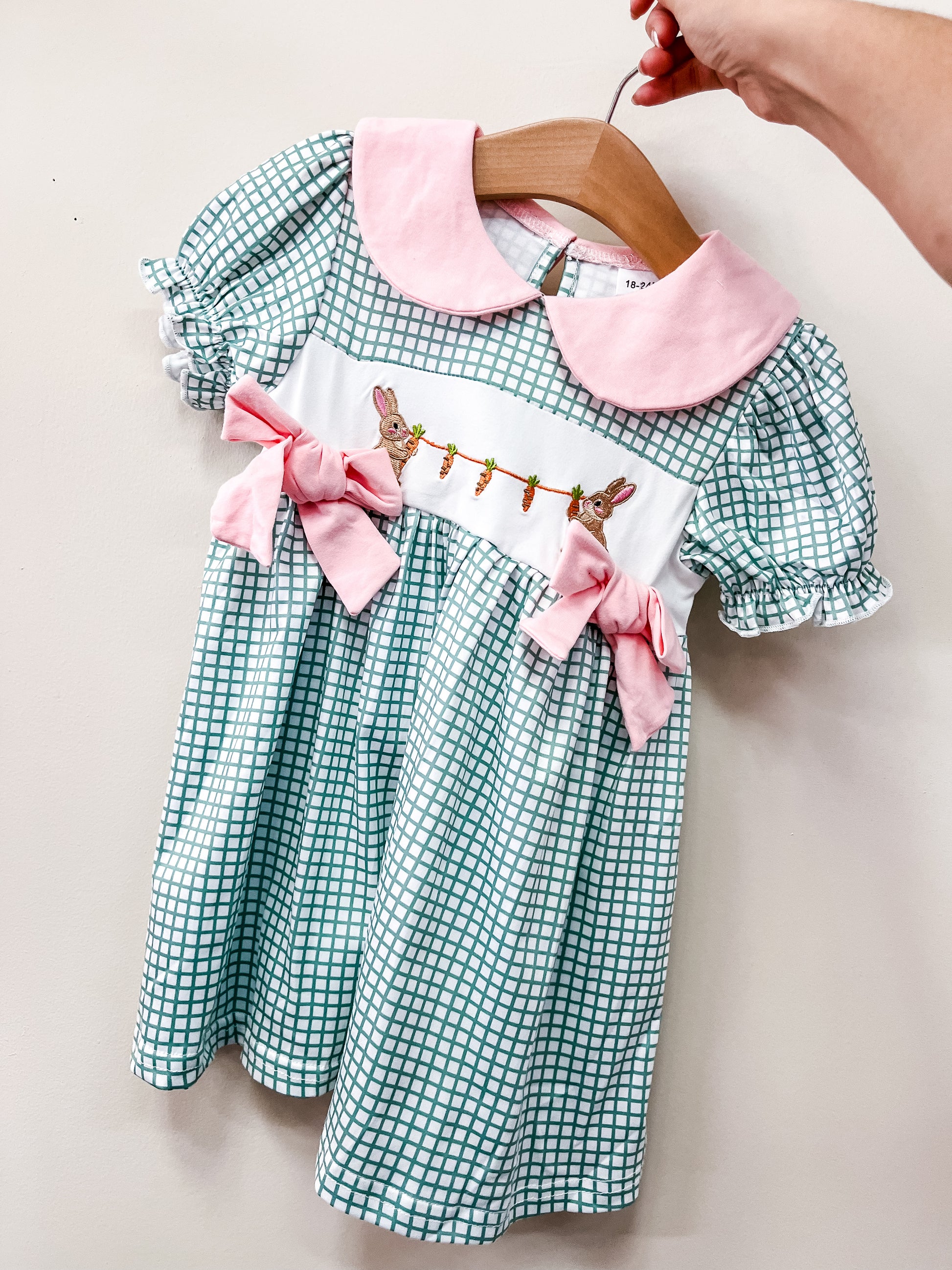 A hand holds the Tiny Tot Bundles Garden Bunny Bow Dress—a green and white checkered child's dress featuring a pink collar, pink bows, and embroidered bunnies and carrots on the white waistband.