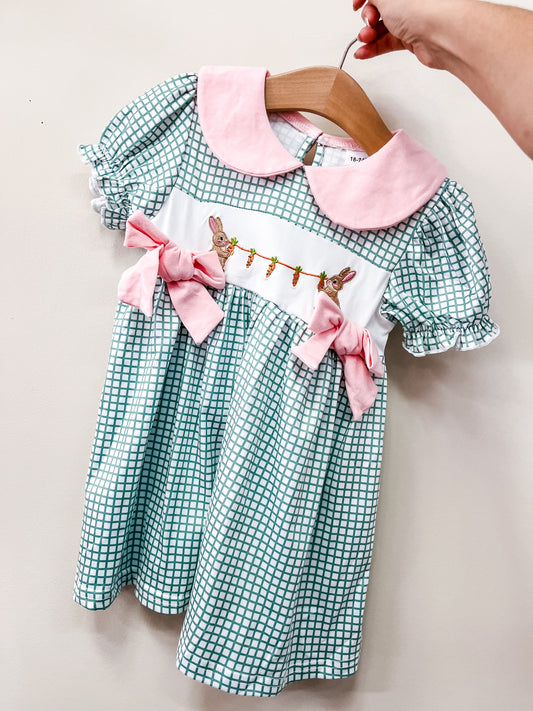 A hand holds the Tiny Tot Bundles Garden Bunny Bow Dress—a green and white checkered child's dress featuring a pink collar, pink bows, and embroidered bunnies and carrots on the white waistband.